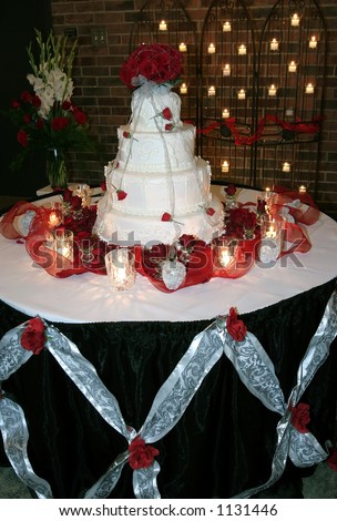 Four tiered wedding cake on table surrounded by rose petals and topped with a rose bouquet.