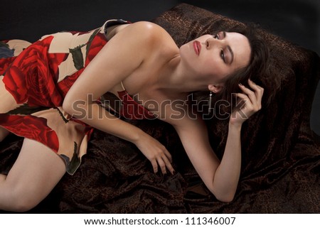 Beautiful young woman in strapless rose-print dress, and red satin high heeled sandals. She gracefully reclines in dramatic pose on dark leopard velvet fabric. Dark background, copy space.