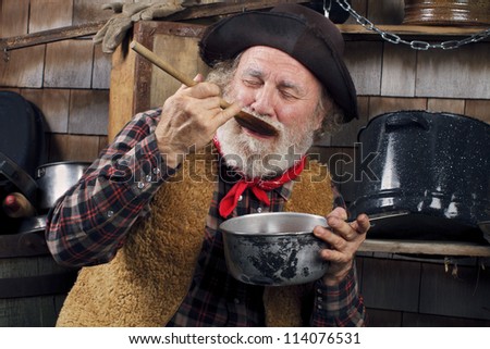 Classic Old West style cowboy with felt hat, grey whiskers, red bandanna. He closes eyes and savors food in a saucepan. Camp cookware and wood shingles in background.