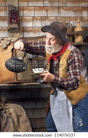 Classic old western style cowboy cook with felt hat, grey whiskers, apron. He is ready to pour tea into a white china tea cup.
