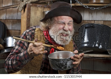 Classic old western style cowboy with felt hat, grey whiskers, red bandanna. He eats beans from a saucepan. Camp cookware and wood shingles in background.