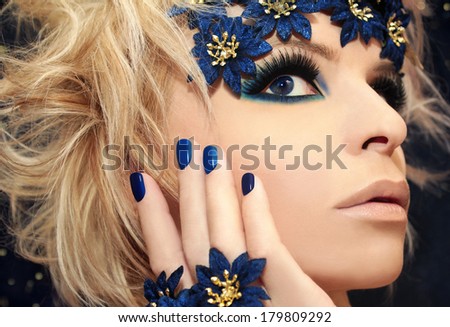 Luxurious blue manicure and makeup on a girl with blond hair and decorative flowers on a dark background.