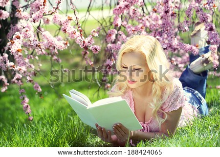 Blonde Girl reading the Book under Cherry Blossom. Beautiful young woman lying on the grass. Outdoor.