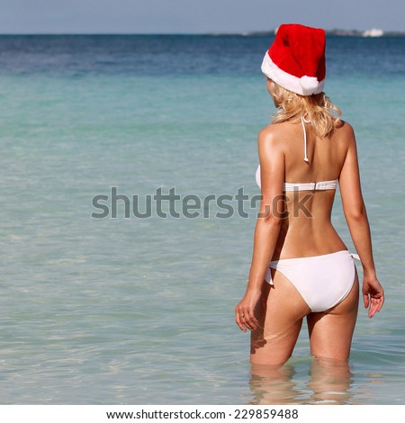 Santa Girl on Tropical Beach. Young woman in Christmas Hat and bikini from behind enjoying blue sea. Vacation.