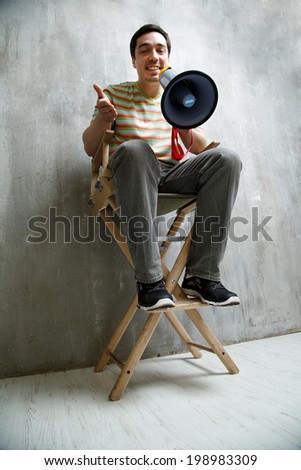 man sitting on a high chair, holding a megaphone and cute smiles on a background of gray textured wall.