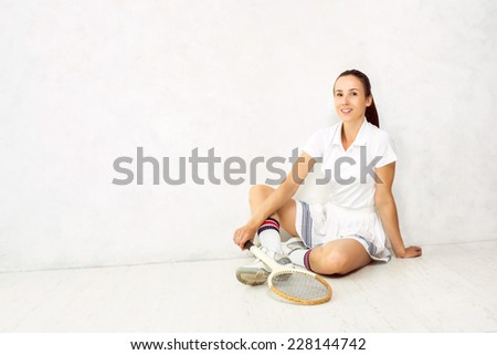 Young beautiful girl with a tennis racket in and in sportswear sitting on a white floor against a white textured wall.