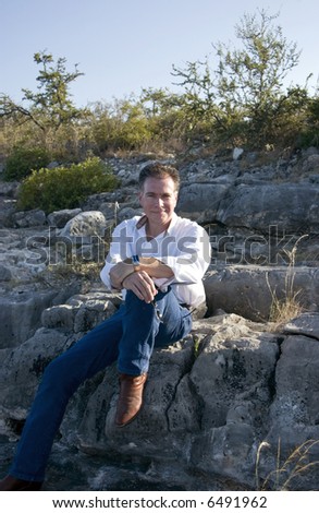 A man, with a friendly smile on his face sitting on a large rock on the side of a hill.