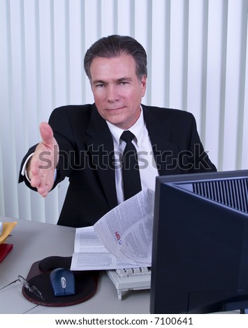 A man sitting at a desk with a legal size document exteding his hand as if to welcome someone.