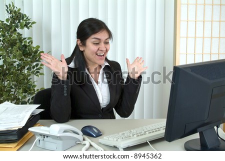 A young woman at her desk, thrilled or surprised by something on her computer monitor.