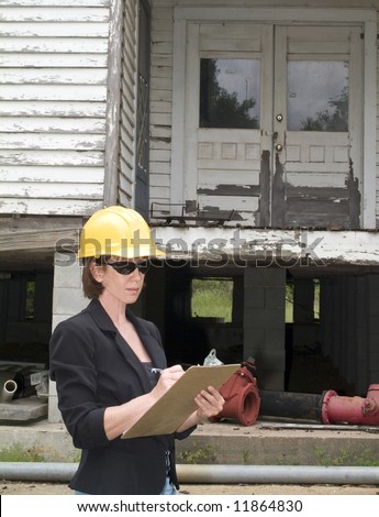 A woman in a hard hat in front of an old rundown house, writing something on a clipboard.