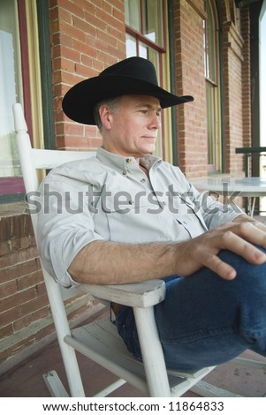A man wearing a cowboy hat, sitting on a porch, in a rocking chair.
