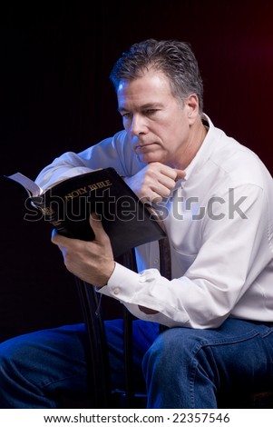 A man sitting astride an old bentwood chair reading a bible, red and blue accent lighting applied.