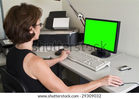 A woman siting at a desk working on her PC with green screen on the flat screen monitor.