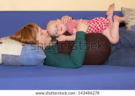 Cute beautiful baby girl playing with her pregnant mother as they rest together on a sofa balancing on her hip kicking her feet in the air