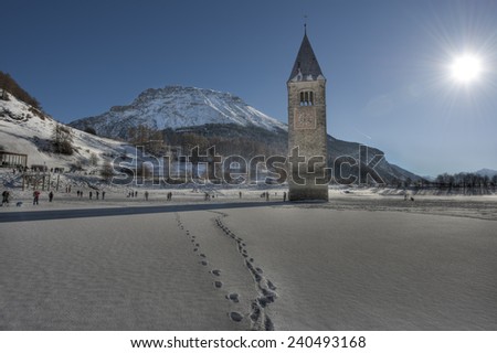 Bell tower in the frozen lake in winter, blue sky with sun, lake Resia, Trentino, Italy