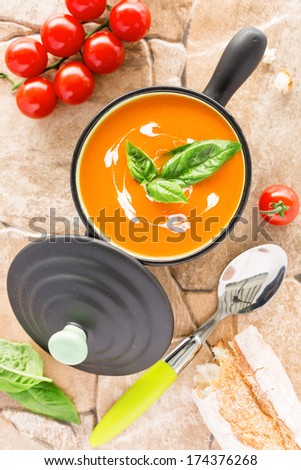 Overhead view of a Tomato soup in a black casserole, drizzled with cream and garnish with leaves of fresh green basil, on orange stone  background