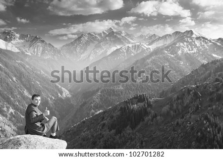 Black and white Image of a young businessman who sits on the top of the mountain and looks at the camera on the background of the beautiful mountains, pleased with the achieved successes.