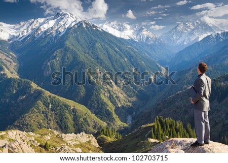 Image of a young businessman with laptop standing at the top of the mountain and looking into the distance to the beautiful mountains, thinking about future plans.