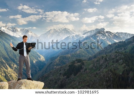 Image of a businessman standing on the top of the mountain, using a laptop with wireless network. In the background a beautiful panorama of the valley with high peaks.