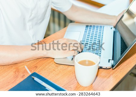 close-up of young man uses laptop at the his workplace, on the table there are Notebook, pen and cup of coffee