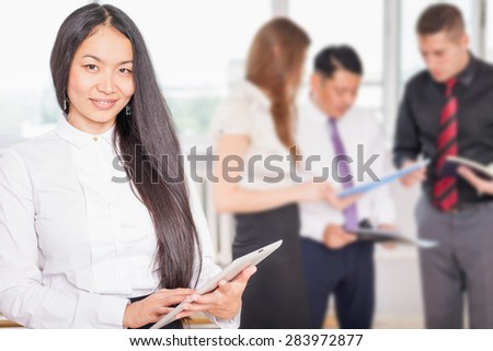Successful asian business woman with magnificent long hair holding tablet PC and business team at background. Image symbolizes corporation or company, uses a new technology for success