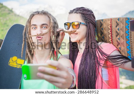 Happy people make selfie on mobile phone and making mustache of their hair, at mountain outdoor. They dressed at hippie style clothing, sunglasses, has long hair, holding skateboard and longboard