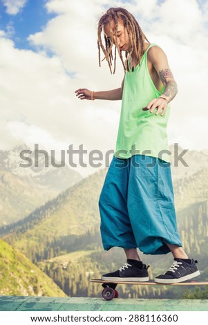 Hippie young and handsome man with skateboard outdoor at mountain. Man riding a skateboard while standing on nose of longboard. He is posing for World Day of skateboarding