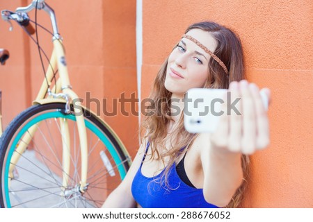 Beautiful and awesome hipster woman make selfie by mobile phone of herself, she sitting near red wall background and vintage city bicycle