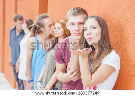 Group of happy young people standing in pairs near wall, they kissing. First couple is sending kiss to camera. Second couple - young man kissing girl cheek. Third couple at background kissing on lips