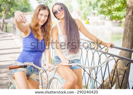 Beautiful sexy women dressed in short shorts travel by city vintage bicycle at summer time. They make selfie on mobile phone