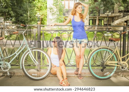 Beautiful young women dressed in shorts resting after travel by city vintage bicycle at heat summer time at park. They are sitting and standing at old bridge at France, Paris