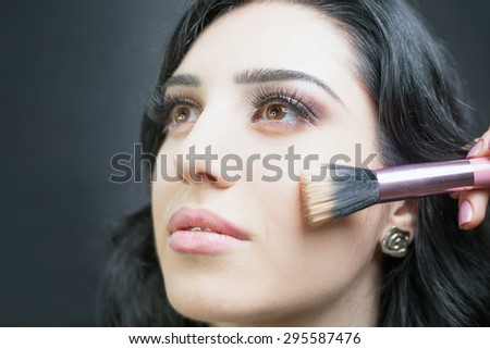 Beautiful woman Arab appearance in the beauty salon with a nice makeup. Make up specialist apply foundation cream on face,     holding in hands a makeup brushe on a dark or black background.