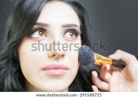 Beautiful woman Arab appearance in the beauty salon with a nice makeup. Make up specialist apply foundation cream on face, holding in hands a makeup brush on a dark or black background.