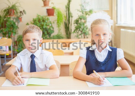 Students or classmates in school classroom sitting together at desk. They are dressed in school uniforms. On table there is a notebook and a pen. The class at background there are plants