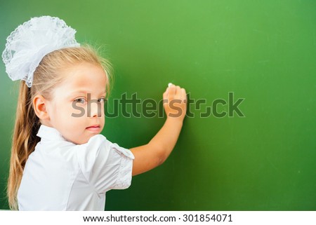 First grade happy schoolgirl wrote on blackboard with chalk at classroom. Little girl dressed in a school uniform. Concept of knowledge day and first of september