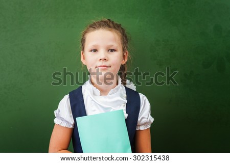 First grade schoolgirl holding a notebook at classroom. Little girl dressed in a school uniform. Concept of knowledge day and first of september. There is a copy space at green blackboard