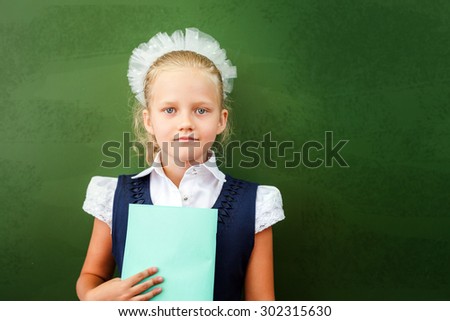 First grade schoolgirl holding a notebook at classroom. Little girl dressed in a school uniform. Concept of knowledge day and first of september. There is a copy space at green blackboard