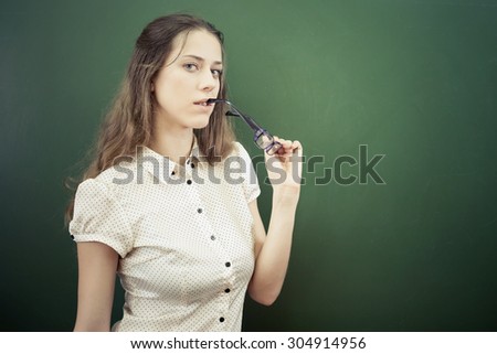 Pretty teacher or student holding glasses near blackboard with at classroom at school or university. She looking at the camera.
