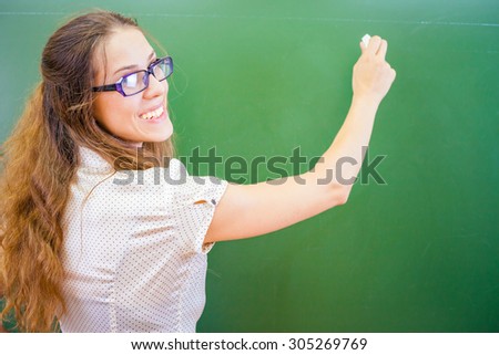 Funny and happy teacher or student wrote on blackboard with chalk at classroom at school or university. She looking at the camera and wearing glasses.