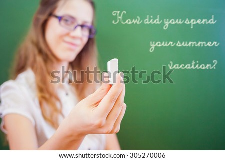 beautiful young woman - teacher or student, holding a chalk near green chalkboard at classroom at university ar school. At background there is a text of How did you spend your summer vacation?