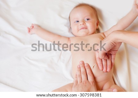 Mother makes massage and stretch for baby stomach, with happy newborn at white bed, apply oil on the hand