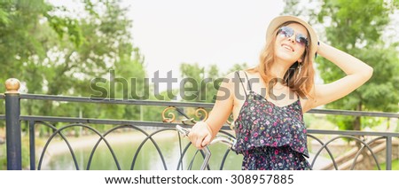Happy young girl traveling, dressed in an elegant summer dress, hat and sunglasses. She stands on the bridge and smiling. There is a copy space. Letterbox format