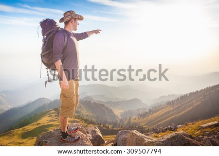 Climbing young adult at the top of summit with aerial view of mountain range. Man is pointing straight
