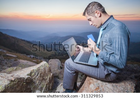 Image of achieve and successful businessman on the top of mountain, using a laptop for online payment by plastic card through the Internet banking