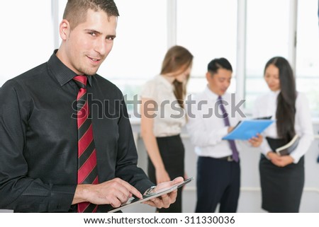 Successful young business man holding tablet pc and business team of women and man at background. Image symbolizes a successful corporation or company, achieve success for intelligent and handsome men