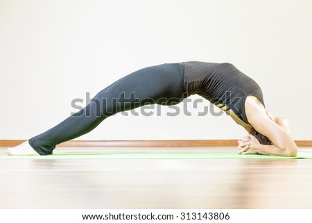 Woman doing exercise of yoga indoor, dressed in black sportswear. She stretching and standing on two hands like bridge pose.