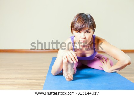 Asian woman stretching at mat before yoga exercise indoor at home. She looking at camera. You can maintain regular workouts at your home for free, be healthy and in good tonus!