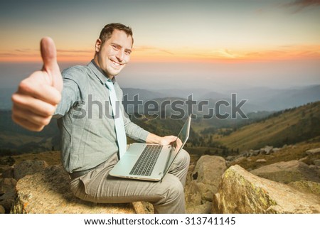 Image of achieve and successful businessman on the top of mountain, using a laptop with thumbs up gesture, looking at camera