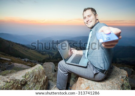 Image of achieve and successful businessman on the top of mountain, using a laptop for online payment by plastic card through the Internet banking. He gives you a credit card