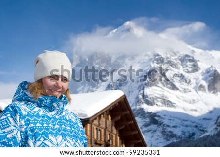 Beautiful smiling young woman looking at the camera. There are a top of alpine mountain summit on the background and a house(hotel). Winter, Switzerland.